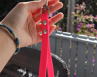 May include: A bright pink dog leash with a silver metal clasp and rivets. The leash is held up against a background of greenery and a wooden fence. The leash is made of a flexible, durable material.