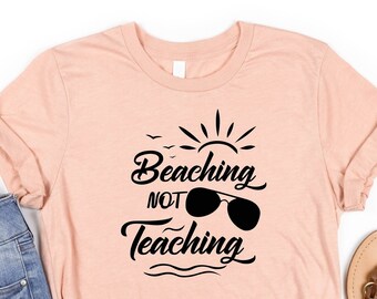 May include: A peach-colored t-shirt with the text "Beaching Not Teaching" in black lettering. The text is stylized with a sun, sunglasses, and waves.