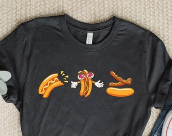 May include: Black t-shirt featuring a cartoon hot dog design. The graphic shows a hot dog breaking apart, a surprised hot dog with arms and eyes, and a hot dog on a bun. The design is in shades of orange, yellow, and brown.