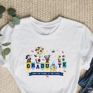 Peut inclure: T-shirt blanc avec un graphique coloré de Mickey Mouse, Minnie Mouse, Dingo, Donald Duck et Pluto portant des chapeaux et des robes de graduation. Le graphique indique "Graduate from the tassel to the castle".