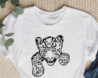 Può includere: Una maglietta bianca con un'illustrazione in bianco e nero di un cucciolo di leopardo che spunta da dietro una superficie. Il cucciolo ha un motivo a macchie e guarda direttamente lo spettatore.