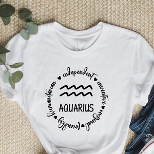 Puede incluir: Camiseta blanca con un gr&aacute;fico circular negro que presenta el signo del zod&iacute;aco Acuario y las palabras "independiente", "inventivo", "humanitario", "amigable" y "original".