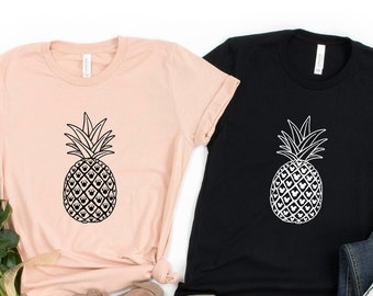 May include: Two t-shirts with a pineapple design. One t-shirt is black and the other is a light pink color. The pineapple design is black on the pink t-shirt and white on the black t-shirt.