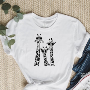 May include: White t-shirt featuring a black graphic of three giraffes wearing sunglasses. The giraffes are standing side by side.