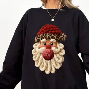 Peut inclure: Sweat-shirt noir avec un motif de Père Noël. Le visage du Père Noël est composé de rouge, beige et imprimé léopard. La barbe est en corde beige et le bonnet rouge avec une bordure léopard. Vêtement de fête.