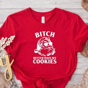 May include: A red t-shirt with a white graphic of Santa Claus wearing sunglasses and the text "Bitch Better Have My Cookies". The shirt is made of a soft material and is suitable for casual wear.