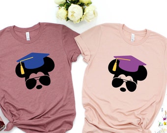 May include: Two t-shirts featuring a black and white silhouette of Mickey Mouse wearing a graduation cap and sunglasses. One shirt is dusty rose in colour and the other is a light pink colour.