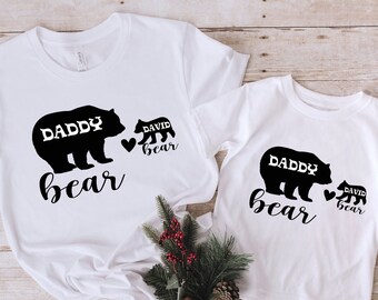May include: Two white t-shirts with black bear silhouettes. The larger shirt says "Daddy bear" and the smaller shirt says "David bear".