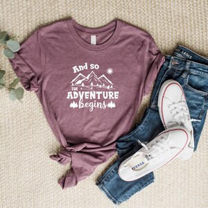 May include: A mauve t-shirt with the text "And so the adventure begins" in white, featuring a mountain range graphic with a sun and trees. The shirt is laid out on a beige surface with a pair of blue jeans and white sneakers.