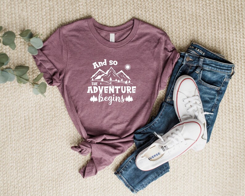 May include: A mauve t-shirt with the text "And so the adventure begins" in white, featuring a mountain range graphic with a sun and trees. The shirt is laid out on a beige surface with a pair of blue jeans and white sneakers.