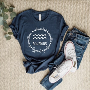 Puede incluir: Una camiseta azul marino con un gr&aacute;fico circular blanco que presenta el s&iacute;mbolo astrol&oacute;gico de Acuario y las palabras "Aquarius", "humanitario", "independiente", "inventivo", "amigable", "inspirador".
