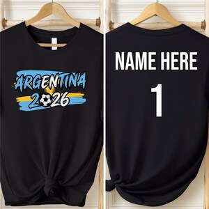 May include: Black t-shirt with the text "ARGENTINA 2026" in blue and yellow, featuring a soccer ball graphic. The back of the shirt displays "NAME HERE" above the number "1" in white.
