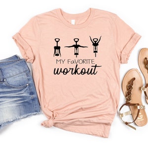 May include: A peach-colored t-shirt with a graphic of three corkscrews in different positions, with the text "MY FAVORITE WORKOUT" below. The t-shirt is paired with denim shorts and brown sandals.