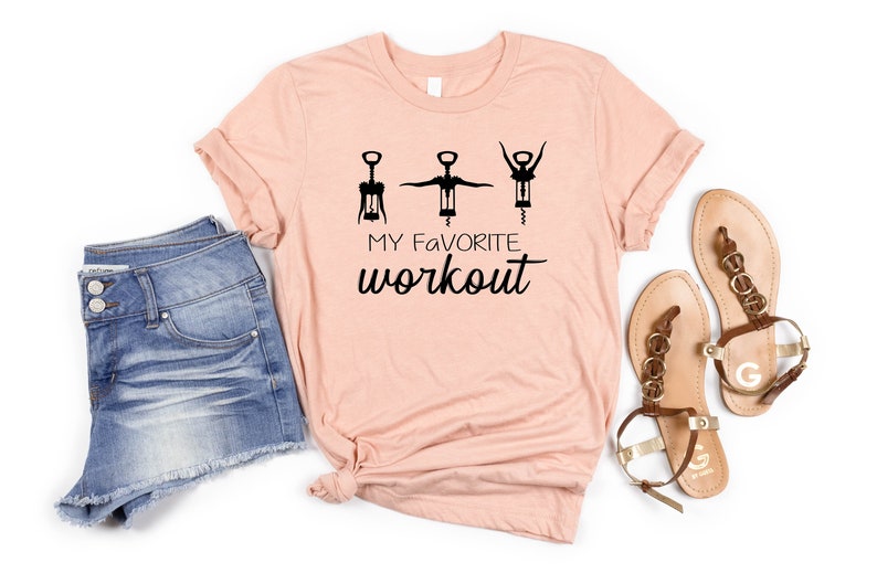 May include: A peach-colored t-shirt with a graphic of three corkscrews in different positions, with the text "MY FAVORITE WORKOUT" below. The t-shirt is paired with denim shorts and brown sandals.
