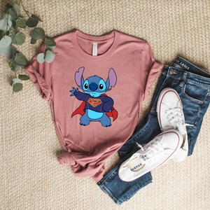 May include: A pink t-shirt with a blue cartoon character wearing a red cape and a Superman logo. The character is Stitch from the Disney movie Lilo & Stitch.