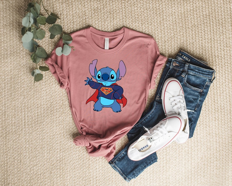 May include: A pink t-shirt with a blue cartoon character wearing a red cape and a Superman logo. The character is Stitch from the Disney movie Lilo & Stitch.
