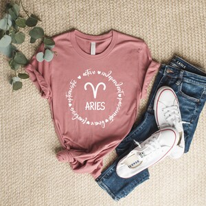 May include: A pink t-shirt with a white circle graphic featuring the zodiac symbol for Aries and the word "Aries" surrounded by the words "optimistic, active, independent, passionate, fearless, brave".