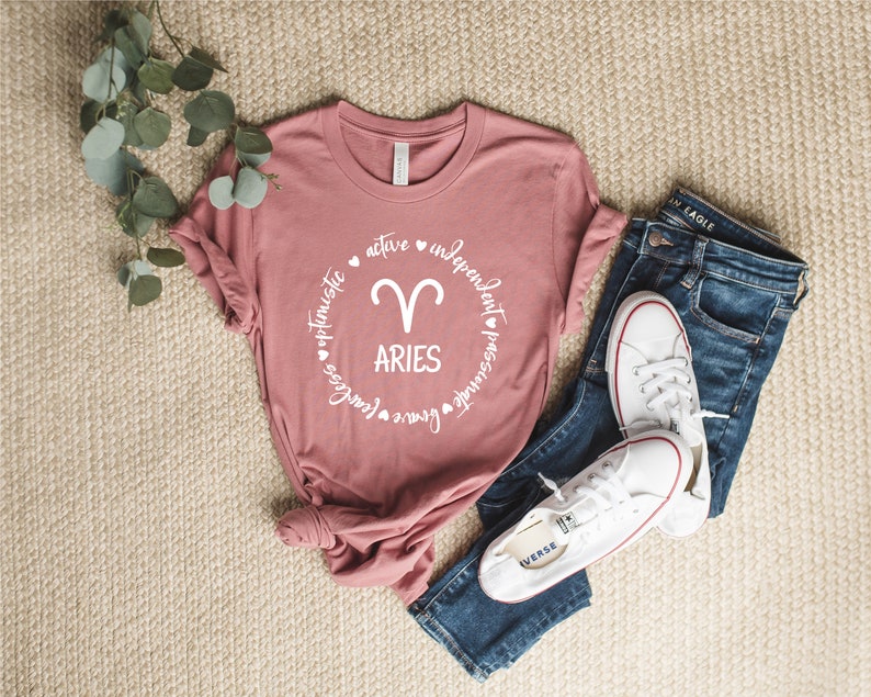 May include: A pink t-shirt with a white circle graphic featuring the zodiac symbol for Aries and the word "Aries" surrounded by the words "optimistic, active, independent, passionate, fearless, brave".