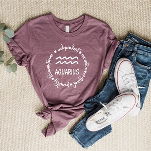 Puede incluir: Una camiseta color malva con un gr&aacute;fico circular blanco que presenta el s&iacute;mbolo astrol&oacute;gico de Acuario y las palabras "Aquarius", "independiente", "humanitario", "inventivo", "fuerte", "amoroso" y "libre".