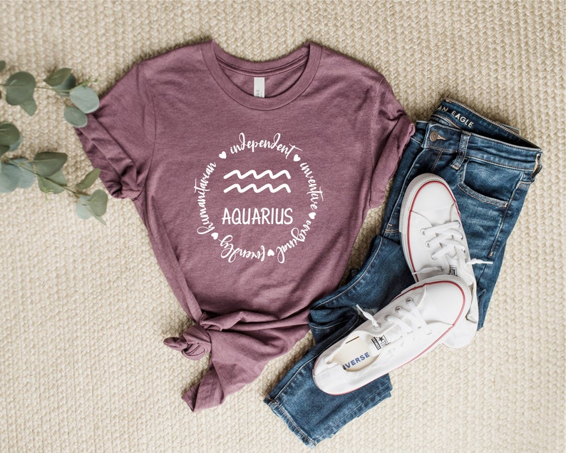 Puede incluir: Una camiseta color malva con un gr&aacute;fico circular blanco que presenta el s&iacute;mbolo astrol&oacute;gico de Acuario y las palabras "Aquarius", "independiente", "humanitario", "inventivo", "fuerte", "amoroso" y "libre".