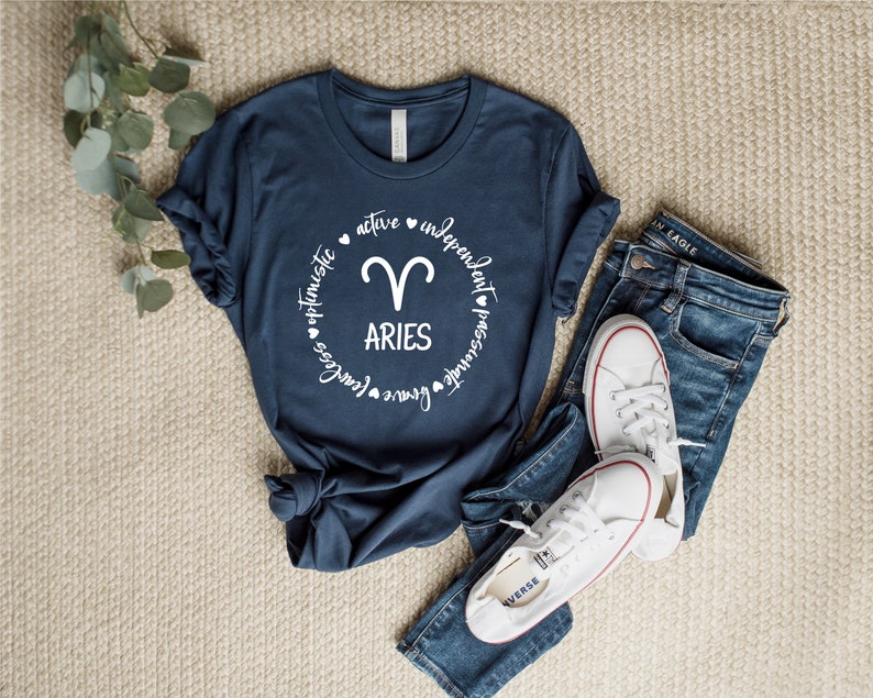 May include: A navy blue t-shirt with a white circle graphic featuring the zodiac symbol for Aries and the text "Aries" surrounded by the words "brave", "love", "fearless", "optimistic", "active", "independent", "passionate", and "powerful".