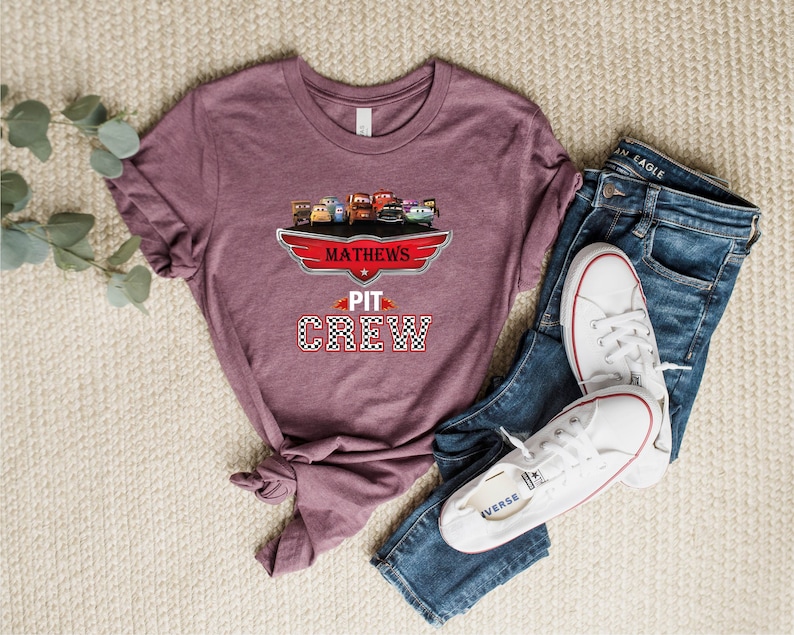 Op de afbeelding: Een mauve t-shirt met een afbeelding van cartoonauto's en de tekst "Mathews' Pit Crew". Het shirt ligt op een beige oppervlak met een paar witte sneakers en een blauwe jeans.
