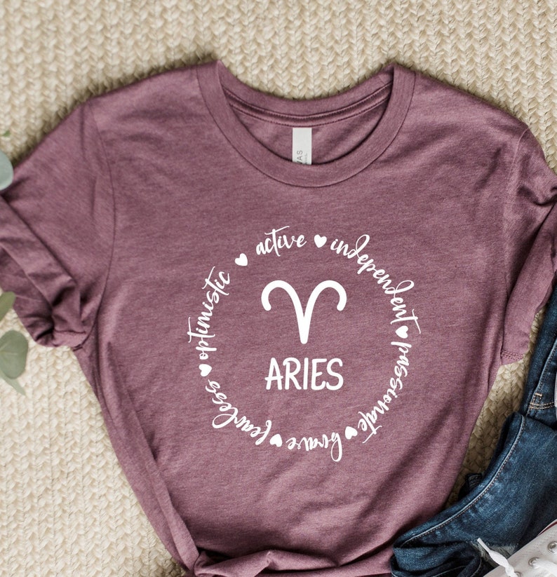 May include: A maroon t-shirt with a white circle graphic featuring the astrological symbol for Aries and the word "ARIES" in white text. The circle is surrounded by the words "active", "independent", "passionate", "brave", "fearless", and "optimistic" in white text.