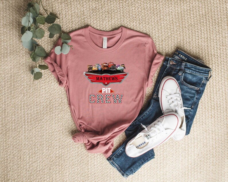 Op de afbeelding: Een roze T-shirt met een afbeelding van cartoonauto's en de tekst "Mathews Pit Crew" in zwart en rood.