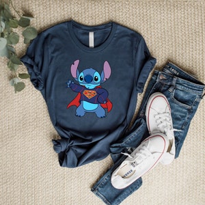May include: A navy blue t-shirt with a cartoon image of Stitch from Lilo & Stitch dressed as Superman. Stitch is wearing a red cape and a yellow Superman logo on his chest.