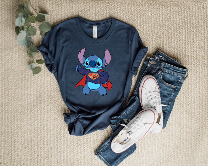 May include: A navy blue t-shirt with a cartoon image of Stitch from Lilo & Stitch dressed as Superman. Stitch is wearing a red cape and a yellow Superman logo on his chest.