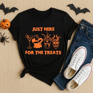 Puede incluir: Camiseta negra con texto y gráficos naranjas de Halloween. El texto dice "JUST HERE FOR THE TREATS" con ilustraciones de un cóctel, bruja, bebida y calavera. La camiseta se combina con vaqueros azules y zapatillas blancas.