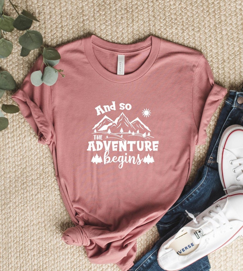 May include: A dusty rose t-shirt with a white graphic that says "And so the adventure begins" with a mountain range and a sun graphic.
