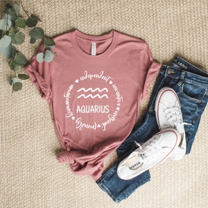 Puede incluir: Una camiseta rosa con un gr&aacute;fico circular blanco que presenta el signo del zod&iacute;aco Acuario y las palabras "humanitario", "independiente", "inventivo", "&uacute;nico", "amoroso" y "adelante".