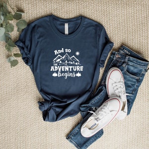 May include: A navy blue t-shirt with white text that reads "And so the adventure begins" with a graphic of mountains and a sun.