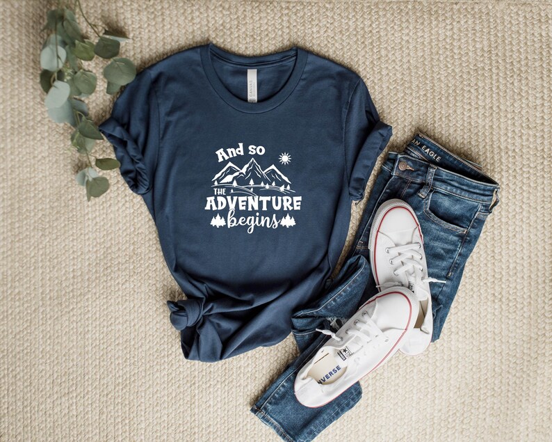 May include: A navy blue t-shirt with white text that reads "And so the adventure begins" with a graphic of mountains and a sun.