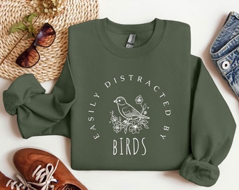 Puede incluir: Una sudadera verde oliva con el texto "EASILY DISTRACTED BY BIRDS" en un diseño circular blanco. El diseño incluye un dibujo lineal de un pájaro posado en flores. La sudadera está doblada, con zapatos marrones y gafas de sol en el fondo.