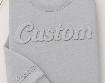 May include: A light gray sweatshirt with the word "Custom" embossed on the front and sleeve. The sweatshirt is folded neatly, with a tag at the neckline. A dandelion seed head is in the upper left corner.