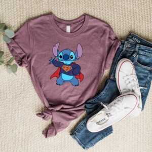 May include: A mauve t-shirt with a blue cartoon character wearing a red cape and a Superman logo. The character is Stitch from Lilo & Stitch.