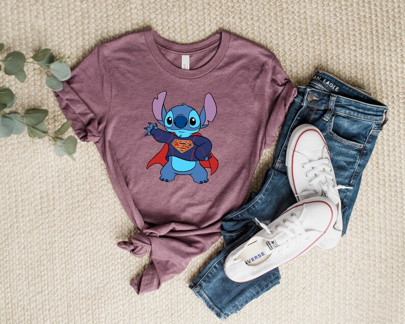 May include: A mauve t-shirt with a blue cartoon character wearing a red cape and a Superman logo. The character is Stitch from Lilo & Stitch.