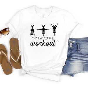 May include: White t-shirt with a graphic of three wine openers and the text "MY FAVORITE workout". The shirt is laid out on a white surface with a pair of brown flip flops, a pair of brown sunglasses, and a pair of blue denim shorts.