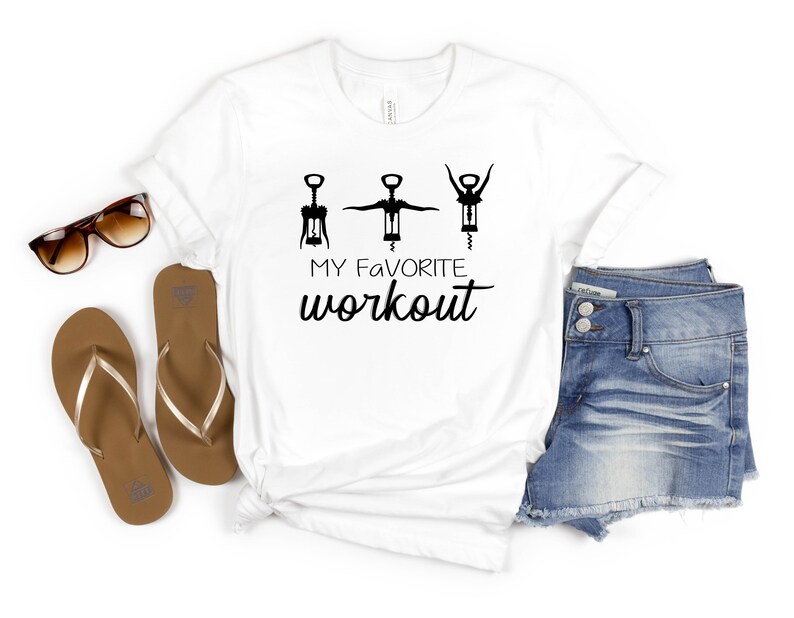 May include: White t-shirt with a graphic of three wine openers and the text "MY FAVORITE workout". The shirt is laid out on a white surface with a pair of brown flip flops, a pair of brown sunglasses, and a pair of blue denim shorts.