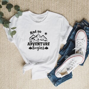May include: White t-shirt with black text that reads "And so the adventure begins" with a mountain and sun graphic.