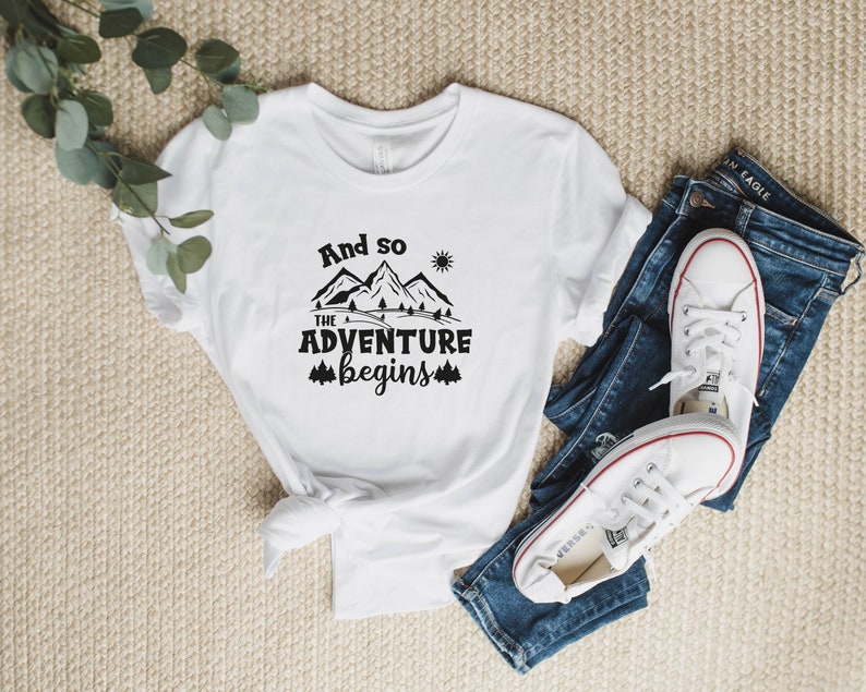 May include: White t-shirt with black text that reads "And so the adventure begins" with a mountain and sun graphic.