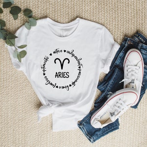 May include: White t-shirt with a black circle graphic featuring the zodiac symbol for Aries and the text "Aries" surrounded by the words "active", "independent", "passionate", "genuine", "fearless", "optimistic".