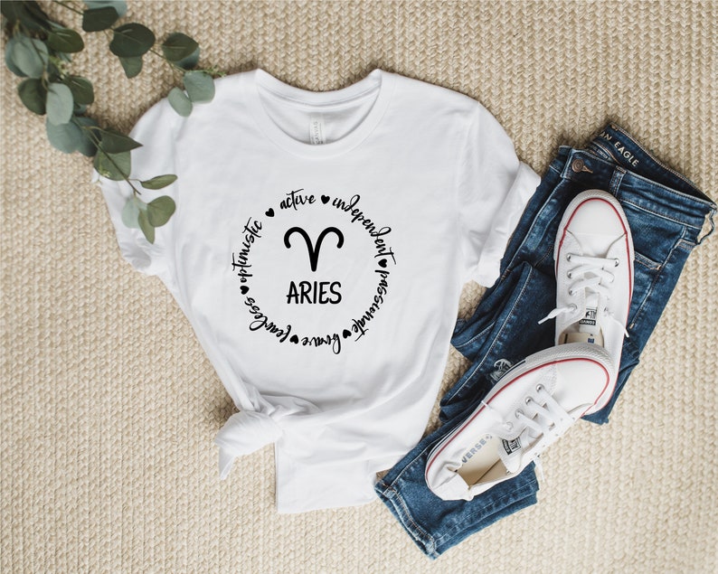 May include: White t-shirt with a black circle graphic featuring the zodiac symbol for Aries and the text "Aries" surrounded by the words "active", "independent", "passionate", "genuine", "fearless", "optimistic".