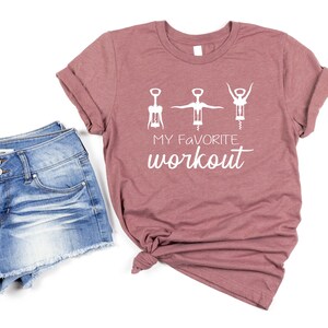 May include: A mauve t-shirt with a white graphic of three wine bottle openers in a workout pose. The text "My Favorite Workout" is printed below the graphic.