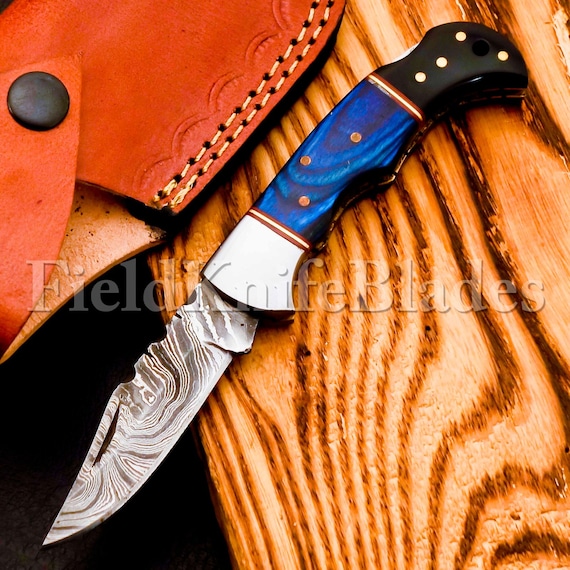 A hand-forged Damascus blade with an olive wood handle and leather sheath — the kind of functional craft object that looks like it belongs in a museum case.