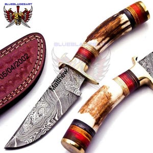 May include: A Damascus steel hunting knife with a brown and white antler handle, red accents, and a leather sheath. The blade is engraved with the name "Matthew". The sheath is embossed with the date "05/04/2002".