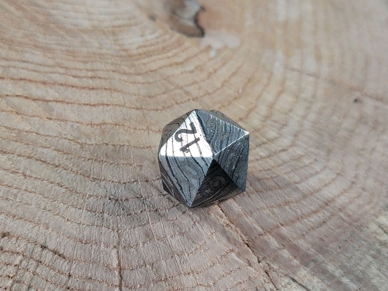 May include: A silver metal 12-sided die with a unique patterned surface. The die is resting on a wooden surface.