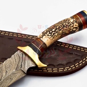Puede incluir: Un cuchillo de caza con una hoja de acero damasco y un mango de asta de ciervo marr&oacute;n con detalles dorados. El cuchillo est&aacute; en una funda de cuero marr&oacute;n con costuras.
